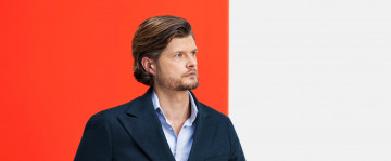 Porträt von Daniel Hiemer, Partner Tax bei Kirkland & Ellis in München, im Dreiviertelprofil mit blauem Sacco und hellblauem Hemd mit geöffnetem Kragen vor orangefarbenem Hintergrund